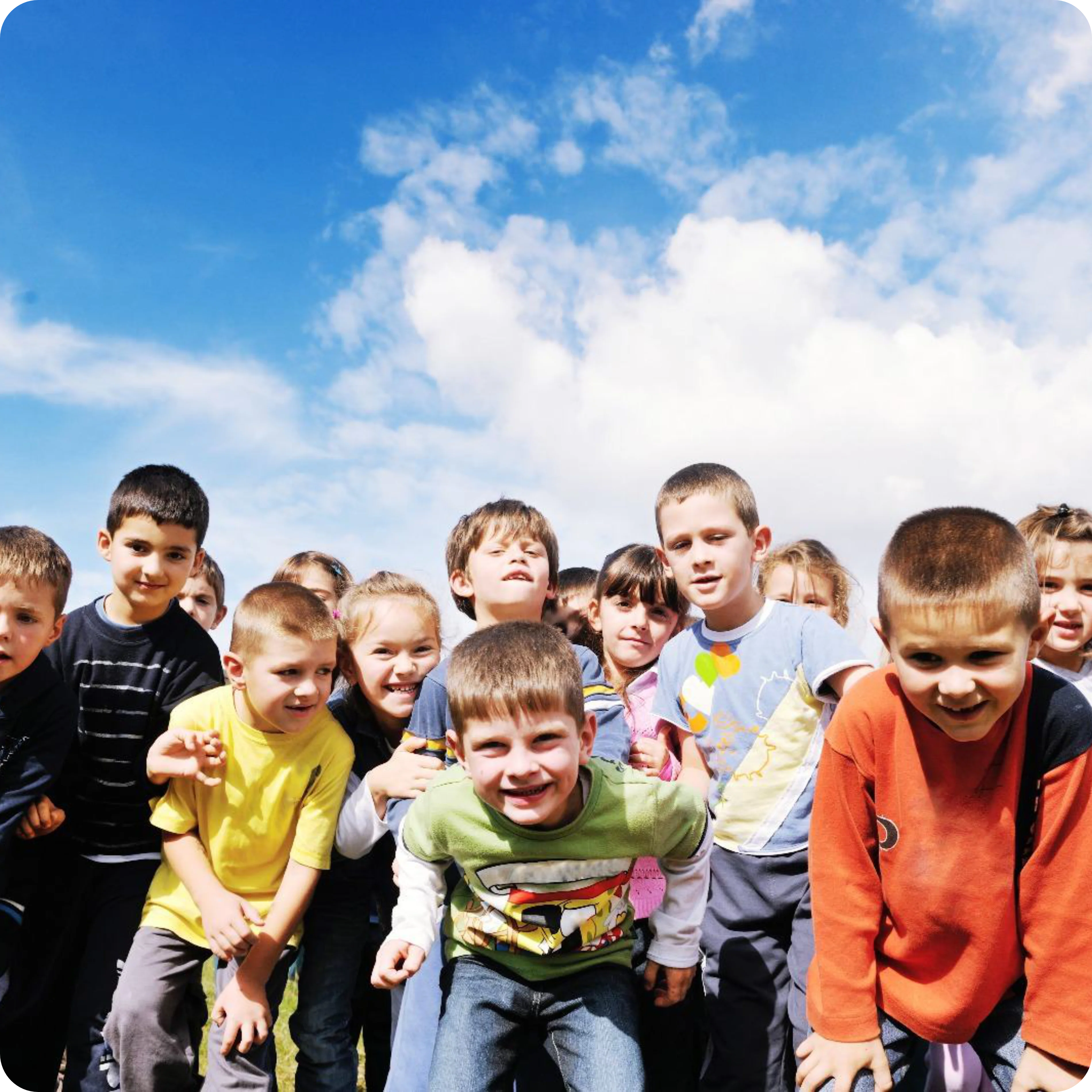 Photo dans un rectangle arrondi représentant une classe d'enfants souriants debout à l'extérieur. Un ciel bleu avec des nuages est visible en arrière-plan.